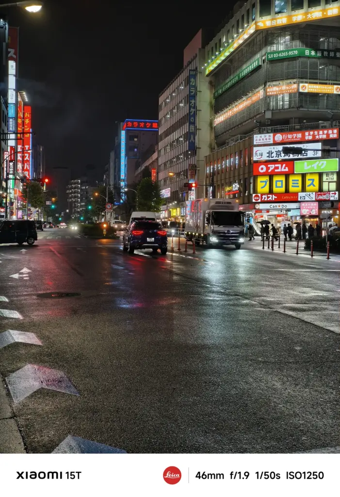 ネオン看板が目立つ雨上がりの街の交差点。看板が光り、トラックや乗用車が走行している。路面の反射が強い。Xiaomi 15T撮影データ: 46mm, f/1.9, 1/50s, ISO1250。