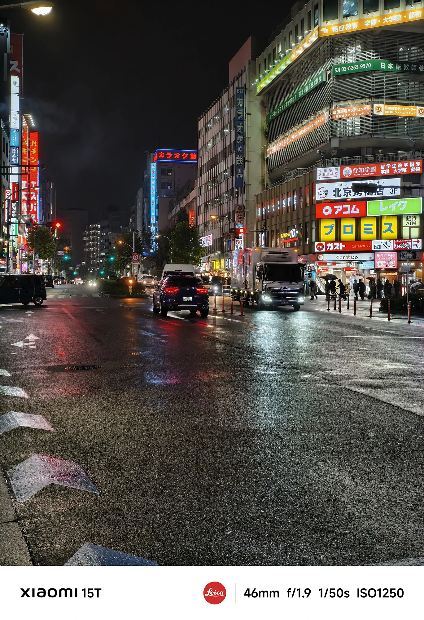 ネオン看板が目立つ雨上がりの街の交差点。看板が光り、トラックや乗用車が走行している。路面の反射が強い。撮影データ: 46mm, f/1.9, 1/50s, ISO1250。