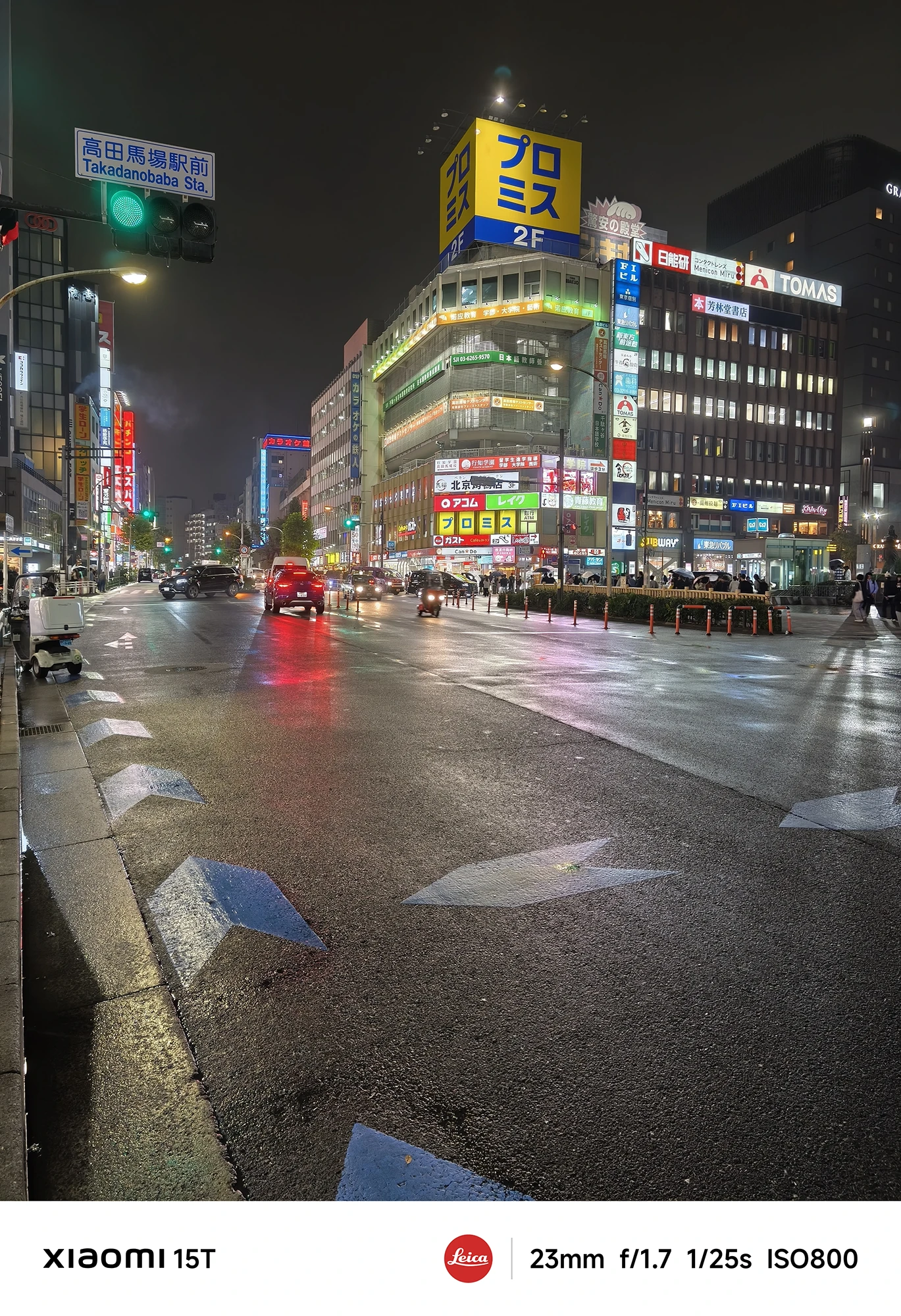 「プロミス」や「トーマス」の看板がある駅前の交差点夜景。赤い乗用車が中央を走行し、濡れた路面に街の光が反射している。撮影データ: 23mm, f/1.7, 1/25s, ISO800。