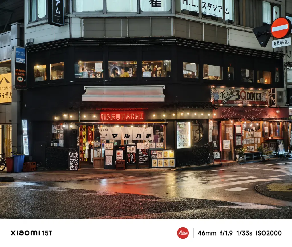 居酒屋「マルハチ」の夜景。黒い建物の1階部分に赤提灯風の装飾と多くのメニュー看板が並び、店内にも人の姿が見える。路面は濡れている。Xiaomi 15T撮影データ: 46mm, f/1.9, 1/33s, ISO2000。