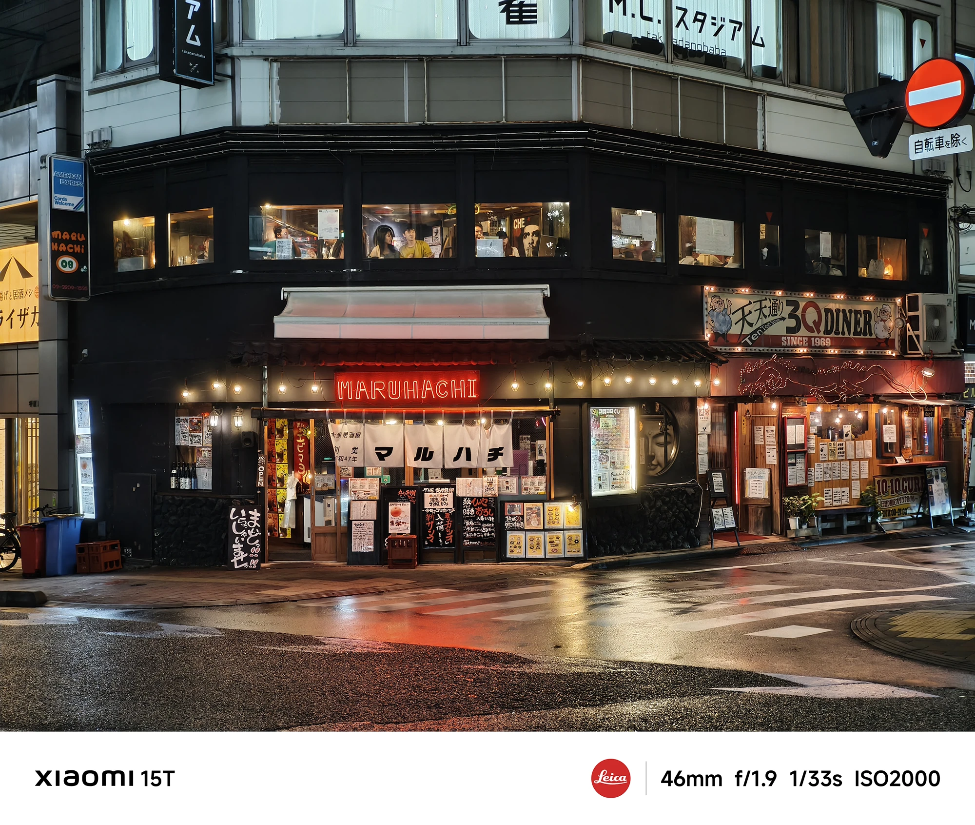 居酒屋「マルハチ」の夜景。黒い建物の1階部分に赤提灯風の装飾と多くのメニュー看板が並び、店内にも人の姿が見える。路面は濡れている。撮影データ: 46mm, f/1.9, 1/33s, ISO2000。