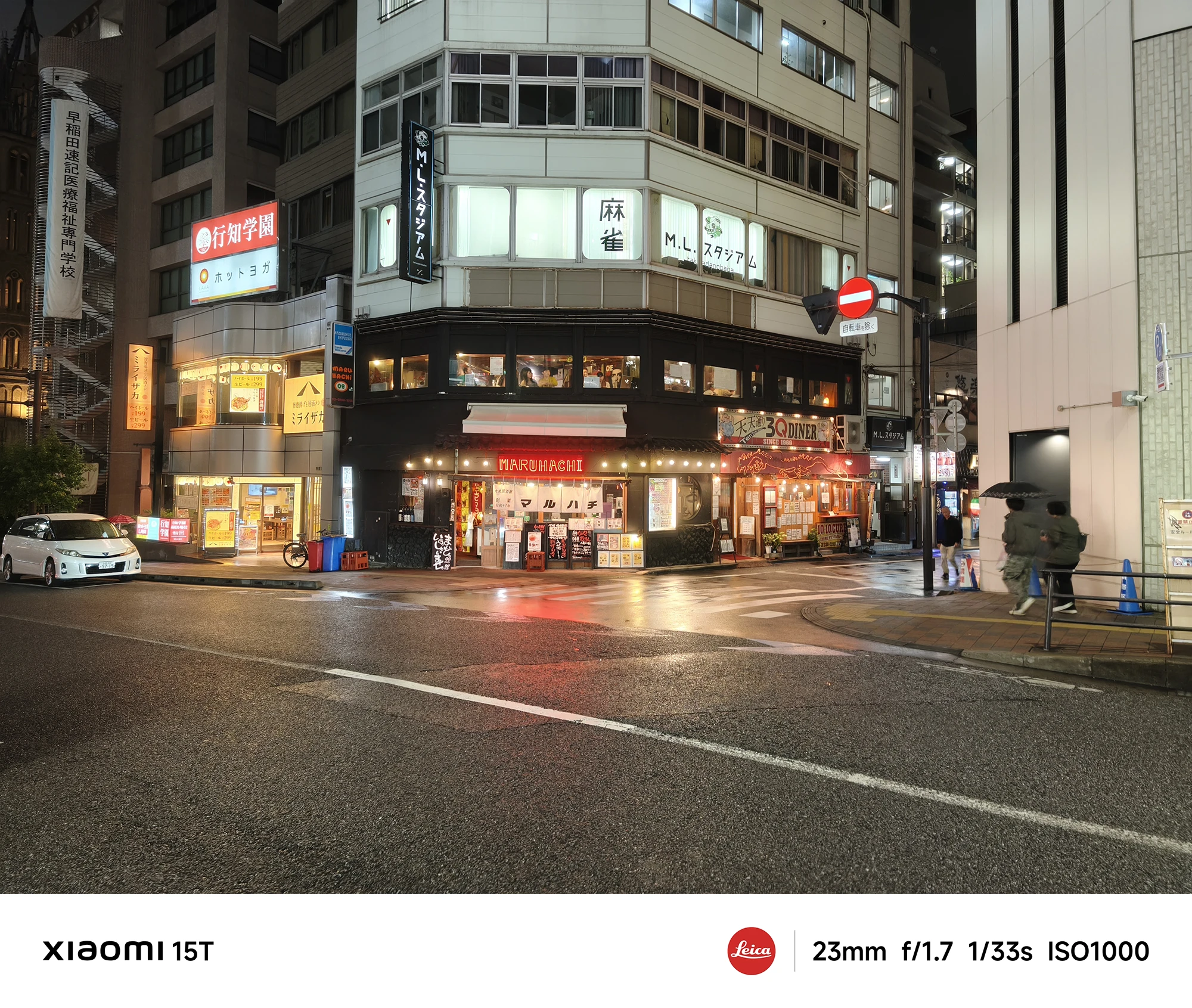 街角にある居酒屋や飲食店が入ったビルの夜景。雨が降る夜の交差点で、白い車が一台停車している。撮影データ: 23mm, f/1.7, 1/33s, ISO1000。