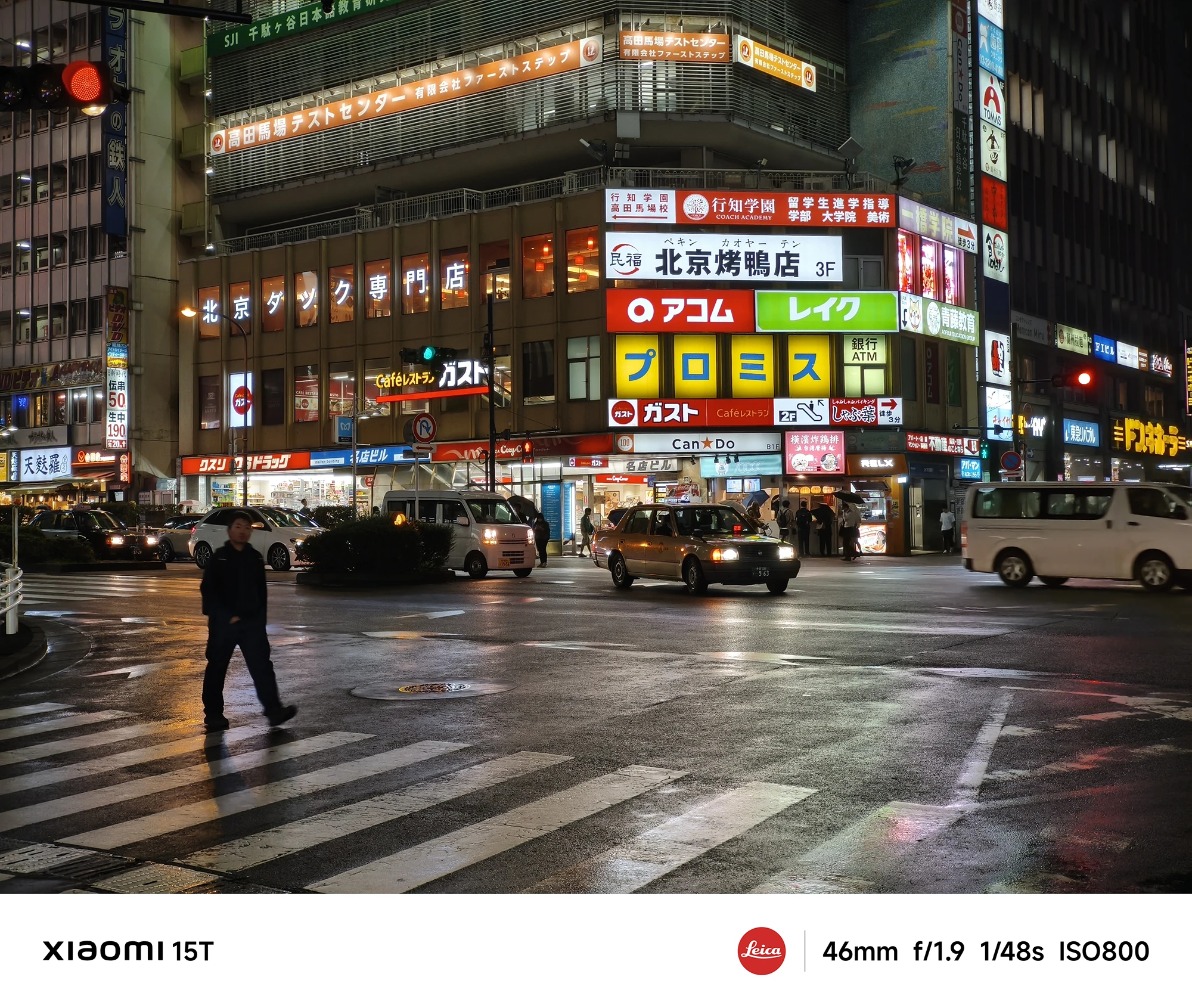 濡れた交差点の夜景。看板が光る雑居ビルの前を、数台の車が走行している。手前には横断歩道を歩く人物のシルエット。撮影データ: 46mm, f/1.9, 1/48s, ISO800。