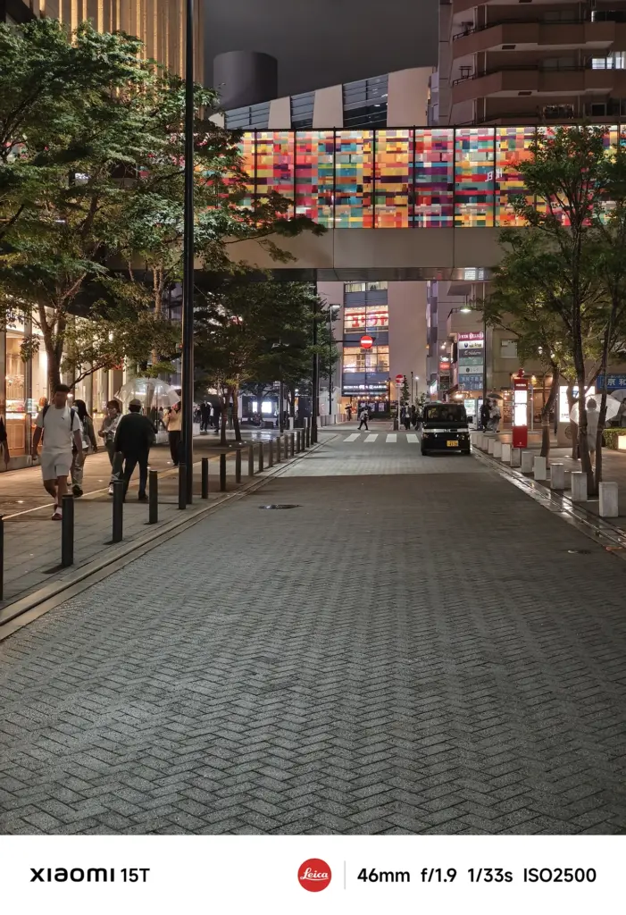 煉瓦敷きの歩行者用道路の夜景。両側に木と建物を配し、奥にカラフルなステンドグラス風の橋が見える。数人が歩いており、黒いタクシーが一台写っている。Xiaomi 15T撮影データ: 46mm, f/1.9, 1/33s, ISO2500。