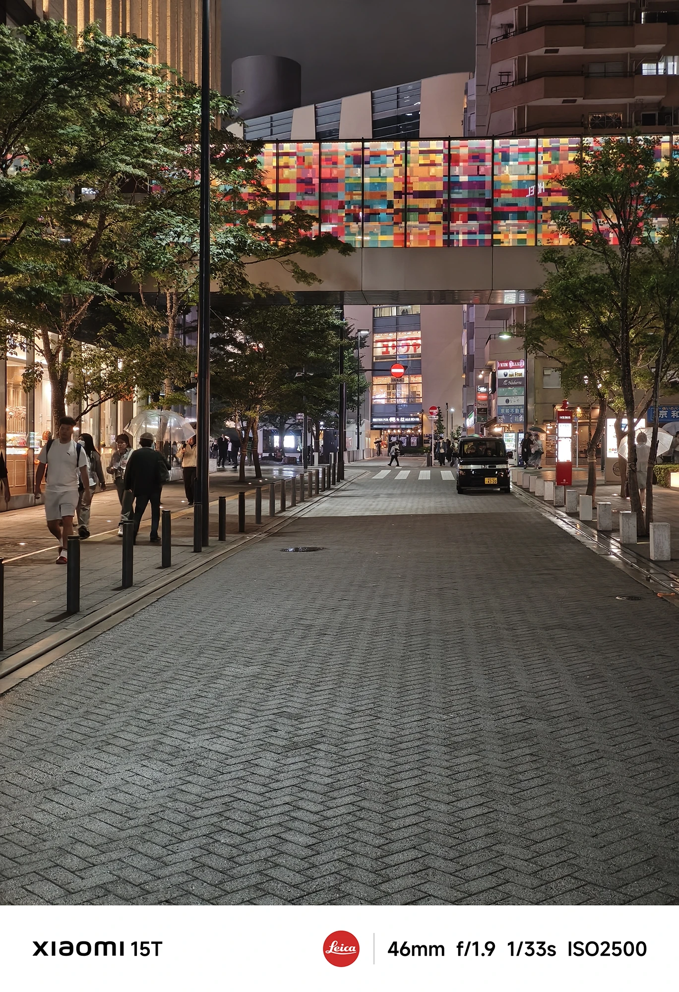煉瓦敷きの歩行者用道路の夜景。両側に木と建物を配し、奥にカラフルなステンドグラス風の橋が見える。数人が歩いており、黒いタクシーが一台写っている。撮影データ: 46mm, f/1.9, 1/33s, ISO2500。