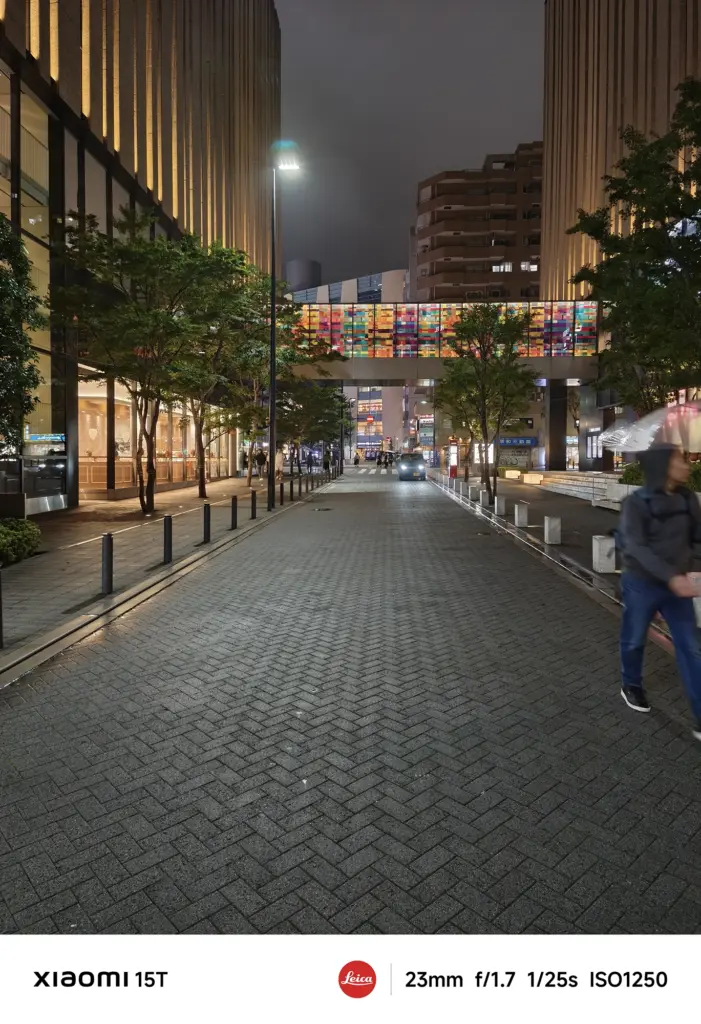 煉瓦敷きの歩道から続く、両脇を建物に挟まれた歩行者用の道。奥にはカラフルなステンドグラス風の橋が見え、右端には傘をさした人物が歩いている。Xiaomi 15T撮影データ: 23mm, f/1.7, 1/25s, ISO1250。