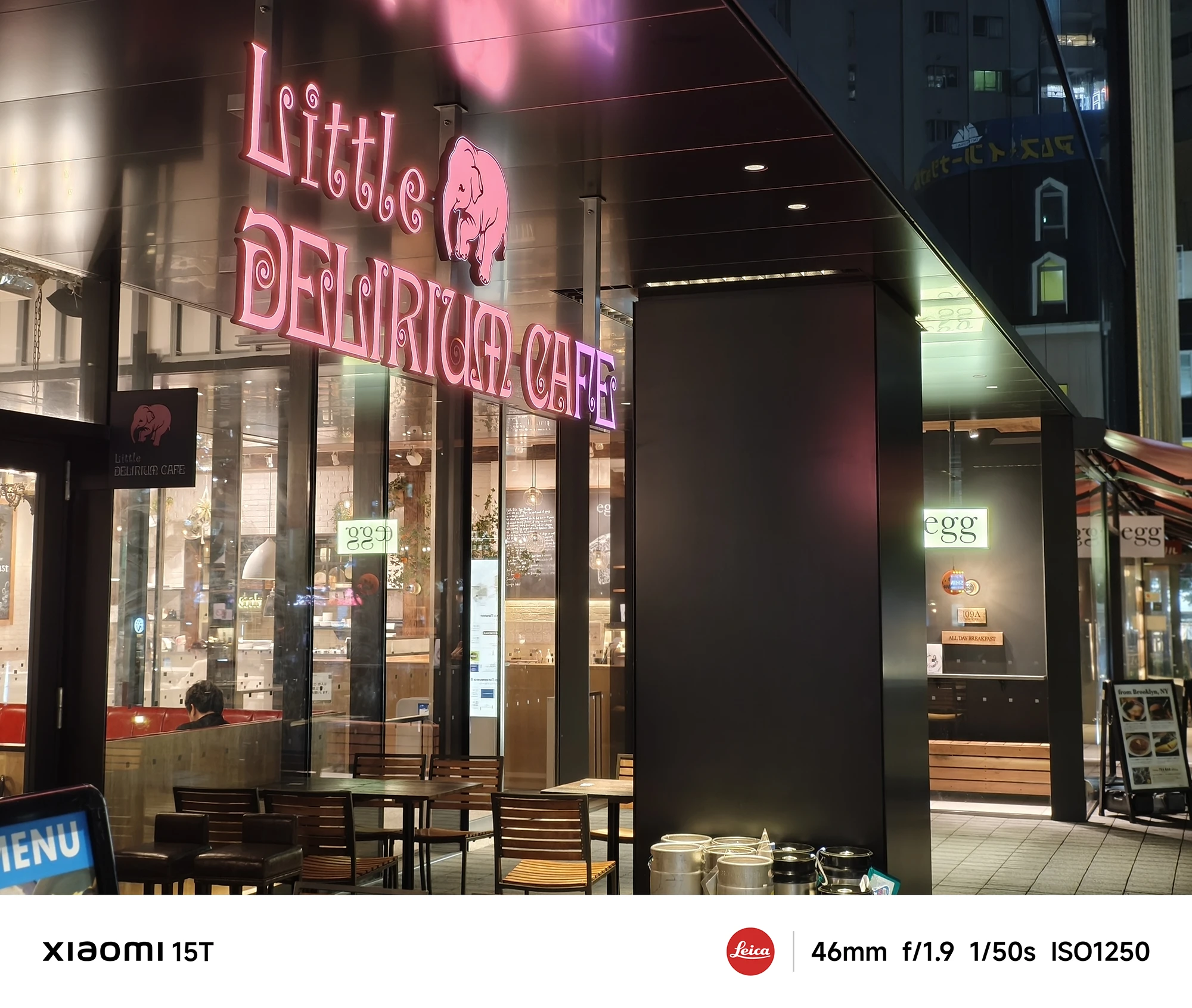 「Little Delirium Cafe」の店の前。ピンク色のネオンサインとテラス席が見える。店内の光が温かく、建物の黒い柱が印象的。撮影データ: 46mm, f/1.9, 1/50s, ISO1250。