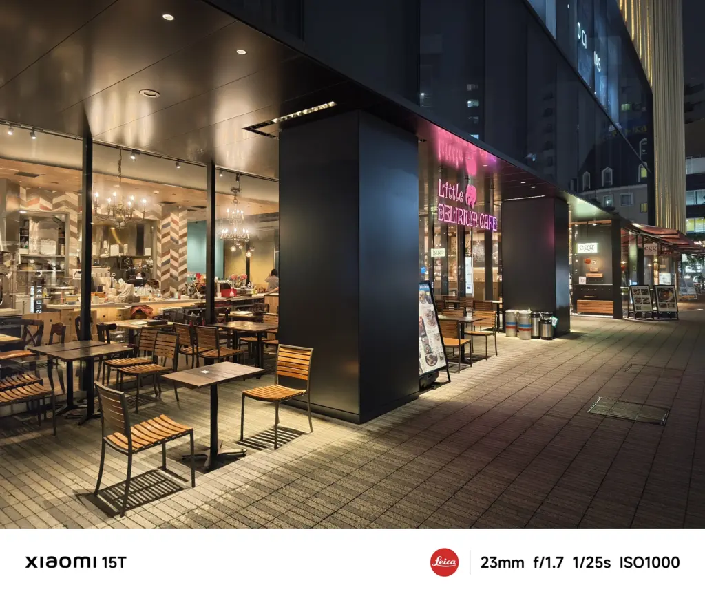 夜の商業ビル1階にあるカフェ「Little Delirium Cafe」のテラス席。木製の椅子とテーブルが並べられ、店内から暖色の光が漏れている。Xiaomi 15T撮影データ: 23mm, f/1.7, 1/25s, ISO1000。