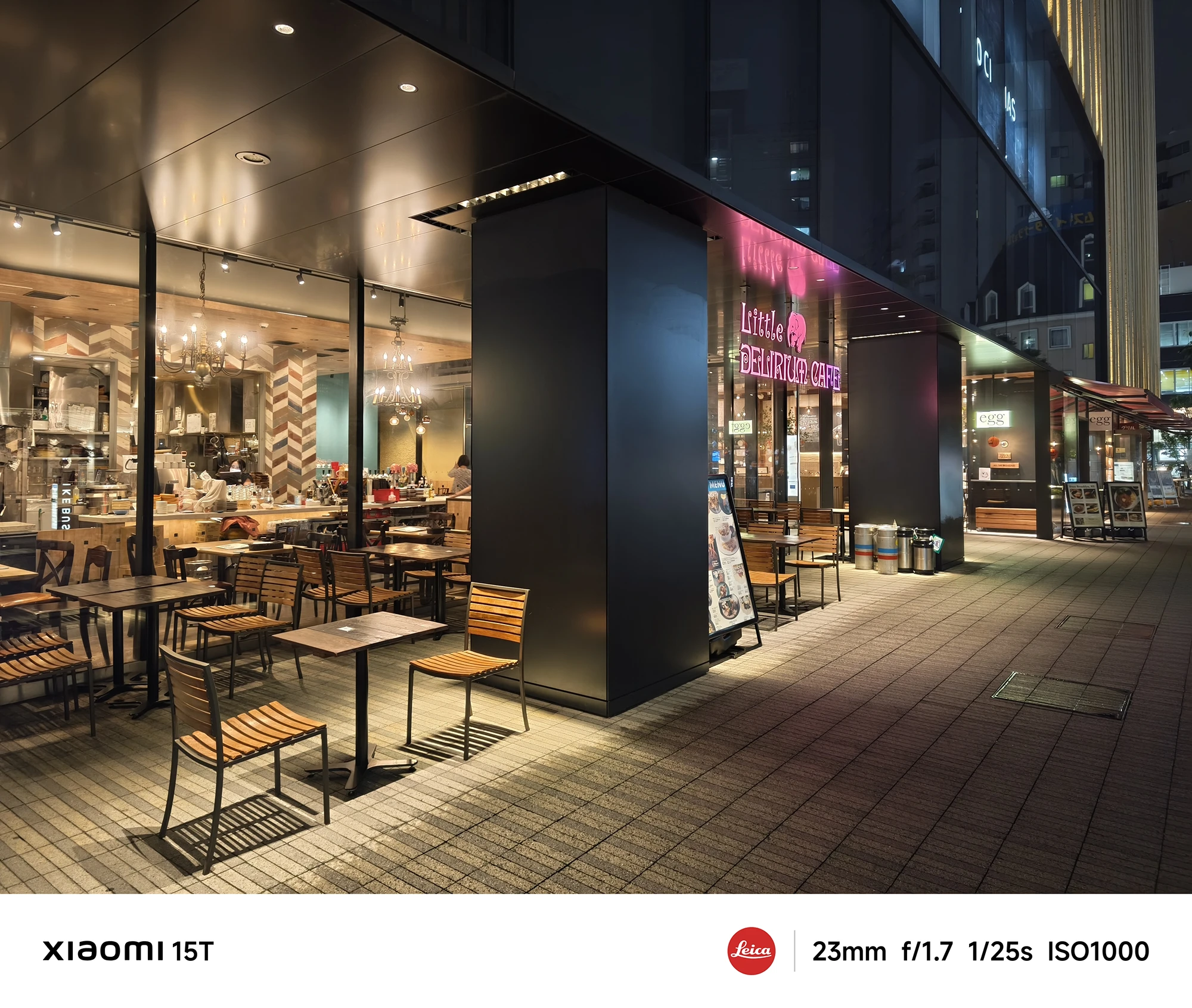 夜の商業ビル1階にあるカフェ「Little Delirium Cafe」のテラス席。木製の椅子とテーブルが並べられ、店内から暖色の光が漏れている。撮影データ: 23mm, f/1.7, 1/25s, ISO1000。
