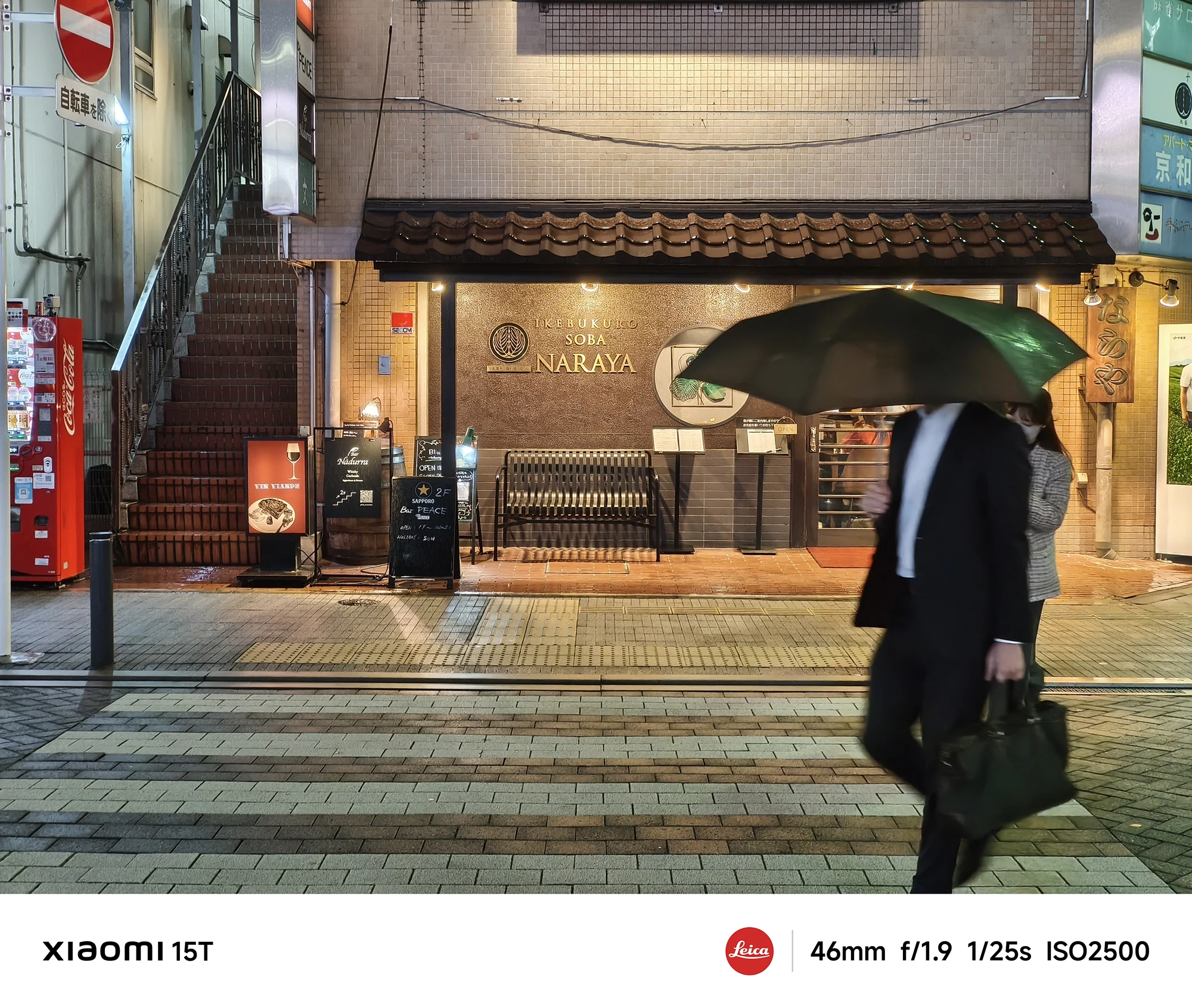 横断歩道を歩くビジネスマン。傘をさし、バッグを持った男性が前景におり、背景には和風の外観のそば店「NARAYA」が見える。撮影データ: 46mm, f/1.9, 1/25s, ISO2500。
