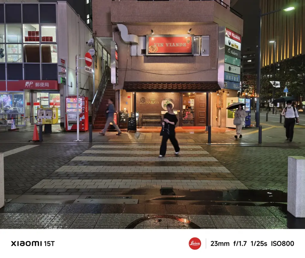 濡れた横断歩道を歩く人々のスナップ。手前に和風居酒屋の建物が見え、周辺は街灯で照らされている。Xiaomi 15T撮影データ: 23mm, f/1.7, 1/25s, ISO800。