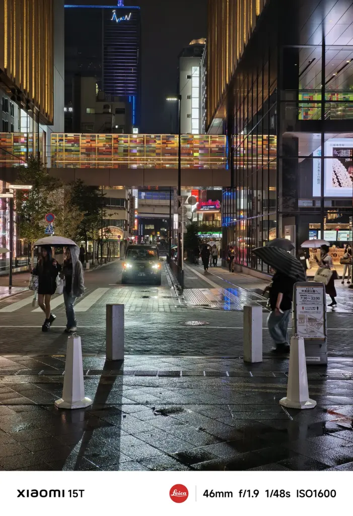 夜の広場と歩行者専用道路。濡れた煉瓦状の地面と奥のカラフルな橋、両側の建物が写っている。傘をさした人々が手前と奥を歩いている。Xiaomi 15T撮影データ: 46mm, f/1.9, 1/48s, ISO1600。