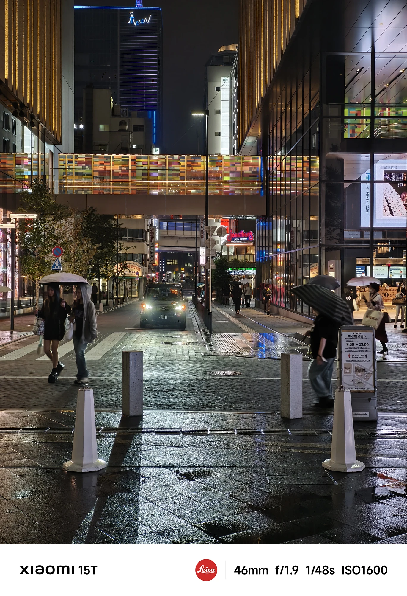 夜の広場と歩行者専用道路。濡れた煉瓦状の地面と奥のカラフルな橋、両側の建物が写っている。傘をさした人々が手前と奥を歩いている。撮影データ: 46mm, f/1.9, 1/48s, ISO1600。