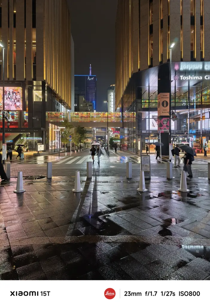 雨に濡れた夜の交差点。両側の高層ビルと奥のタワーがライトアップされ、傘をさした数人が写っている。手前の濡れた路面には建物の光が反射している。Xiaomi 15T撮影データ: 23mm, f/1.7, 1/27s, ISO800。
