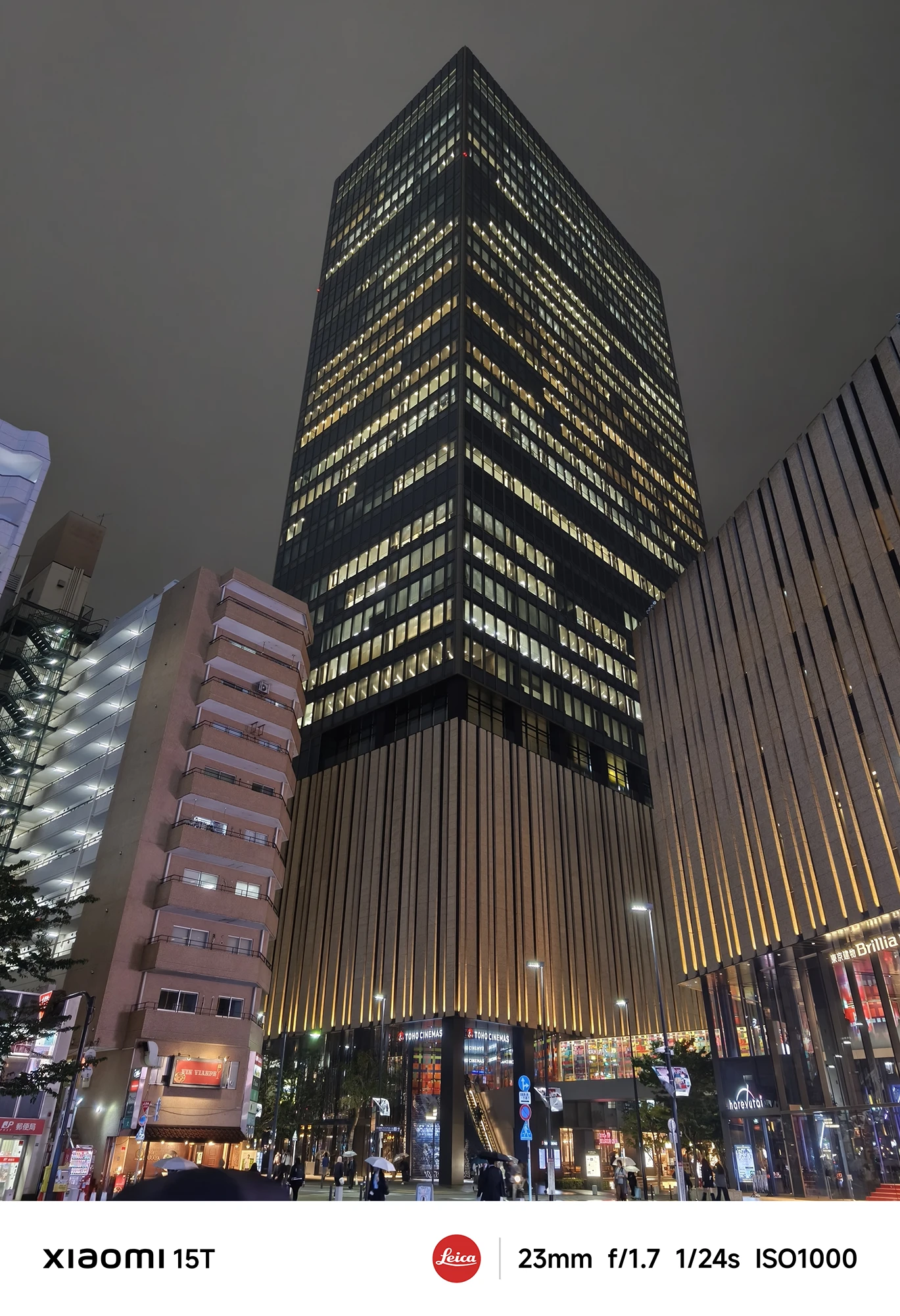都会の夜空に向かってそびえ立つ高層ビルの下からのアングル。低層階は木目の意匠でライトアップされ、多くの窓に灯りが点っている。撮影データ: 23mm, f/1.7, 1/24s, ISO1000。