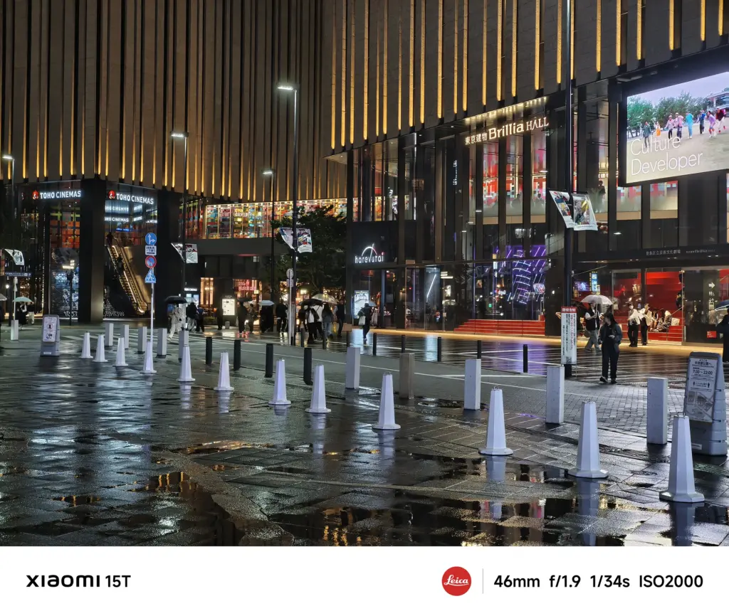 商業施設の広場の夜景。「TOHO CINEMAS」「Brillia HALL」などが入った建物が並び、濡れた広場には白い円錐形のコーンが点々と置かれている。数人が傘をさして歩いている。Xiaomi 15T撮影データ: 46mm, f/1.9, 1/34s, ISO2000。