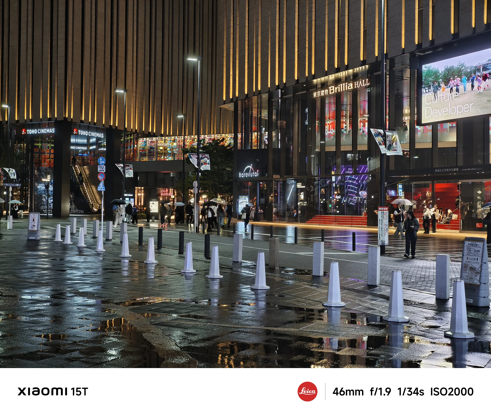 商業施設の広場の夜景。「TOHO CINEMAS」「Brillia HALL」などが入った建物が並び、濡れた広場には白い円錐形のコーンが点々と置かれている。数人が傘をさして歩いている。撮影データ: 46mm, f/1.9, 1/34s, ISO2000。