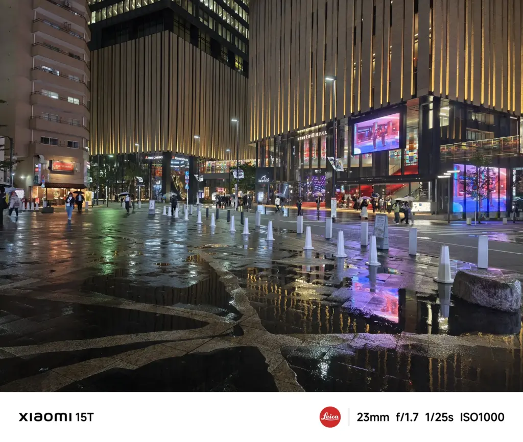 商業施設に囲まれた広場の雨の夜景。濡れた地面の大きな水たまりに周囲の光や建物のラインが反射し、数人が傘をさして歩いている。Xiaomi 15T撮影データ: 23mm, f/1.7, 1/25s, ISO1000。