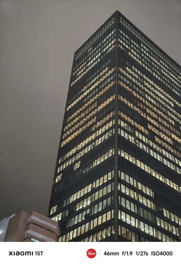 夜空にそびえ立つ高層ビルのクローズアップ。ほぼ全面が窓の巨大な建物の窓の多くに室内の光が灯っている。空は暗い。Xiaomi 15T撮影データ: 46mm, f/1.9, 1/27s, ISO4000。