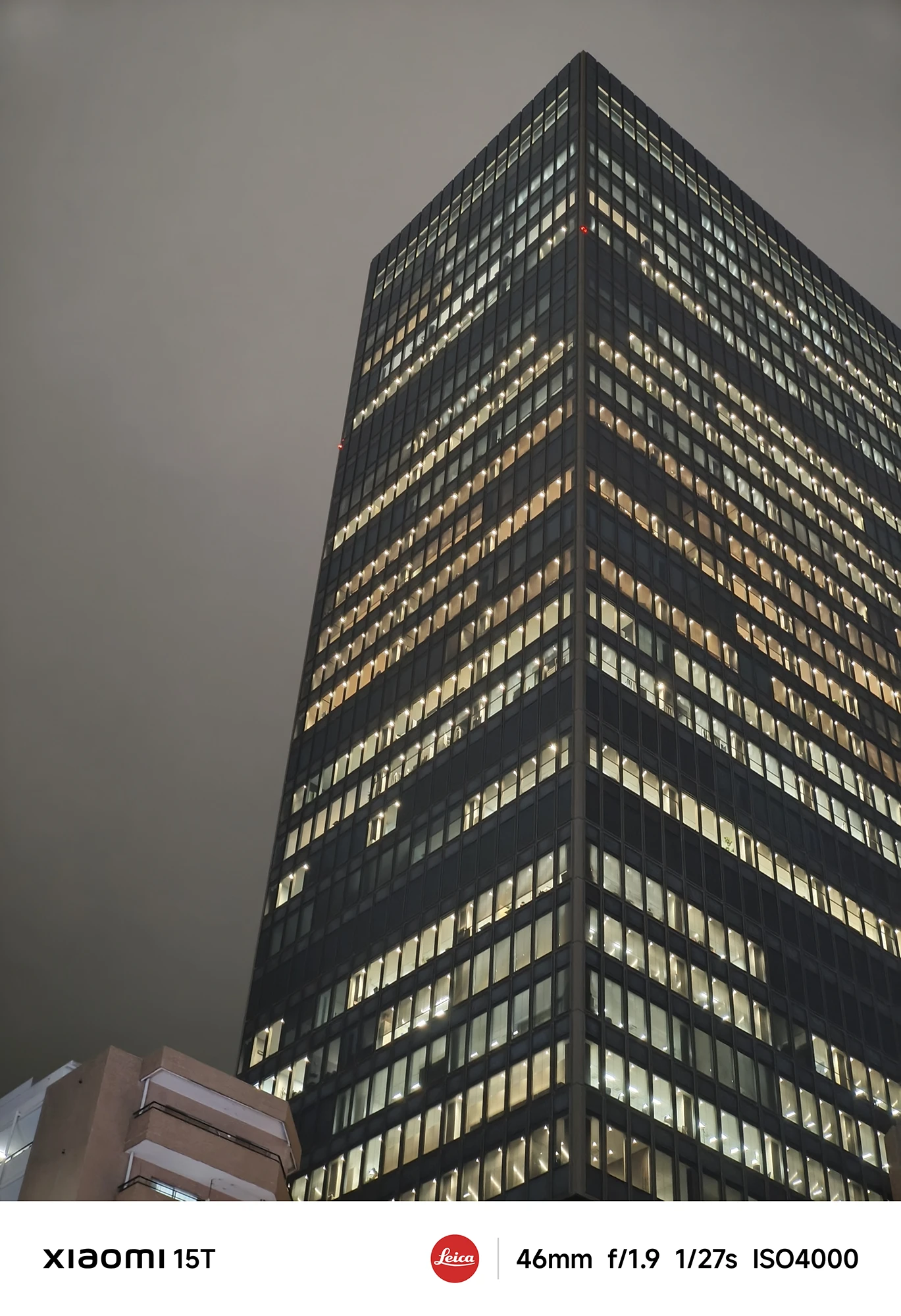 夜空にそびえ立つ高層ビルのクローズアップ。ほぼ全面が窓の巨大な建物の窓の多くに室内の光が灯っている。空は暗い。撮影データ: 46mm, f/1.9, 1/27s, ISO4000。