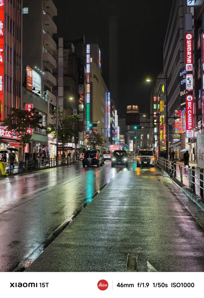 ネオンが輝く繁華街のメインストリート。赤や緑の看板が並び、濡れたアスファルトの道路を数台の車が走行している。濡れた路面が街の光を反射している。Xiaomi 15T撮影データ: 46mm, f/1.9, 1/50s, ISO1000。