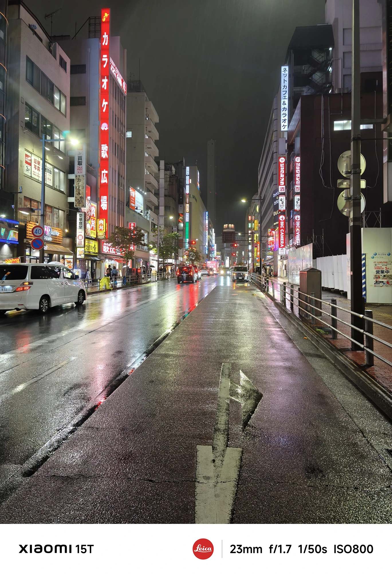 赤いネオンが光るカラオケ店などの看板が並ぶ、雨の夜の繁華街の通り。濡れた路面が車の白いヘッドライトや赤いテールランプを反射している。撮影データ: 23mm, f/1.7, 1/50s, ISO800。