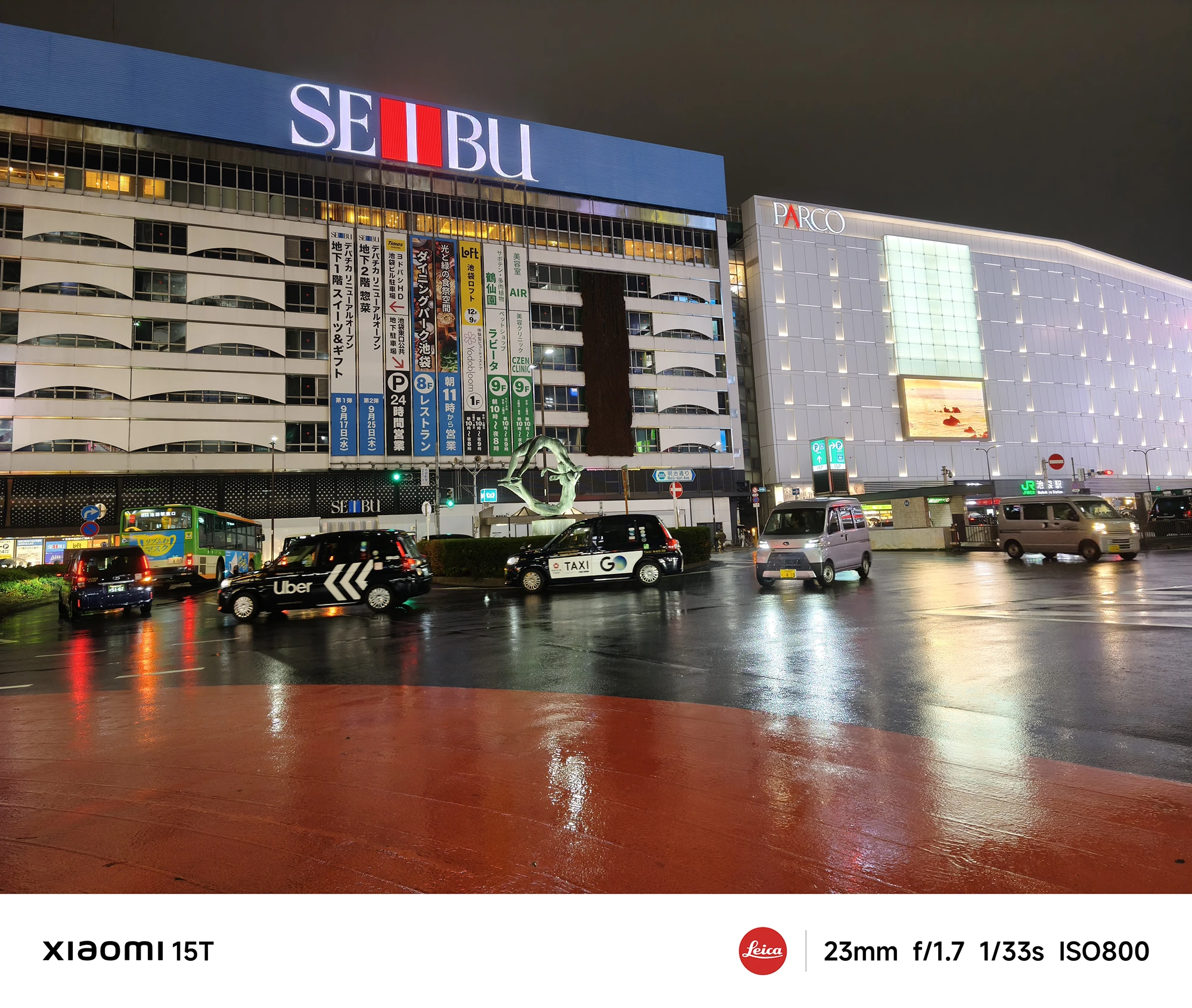 「SEIBU」や「PARCO」の建物が見える、雨上がりの広めの交差点。Uberやタクシーのラッピングがされた車を含め、数台の車が停車している。撮影データ: 23mm, f/1.7, 1/33s, ISO800。