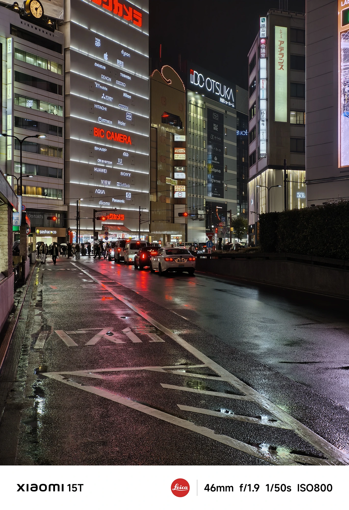 夜の繁華街の通り。「BIC CAMERA」のロゴと家電メーカーの看板が並ぶ建物の前を、テールランプを光らせた車が走行している。濡れた路面がネオンを反射している。撮影データ: 46mm, f/1.9, 1/50s, ISO800。