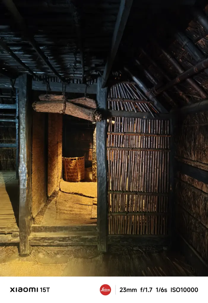 暗い屋内の写真。竹や藁、木の柱でできた日本の古民家のような内部構造で、奥の通路が暖色系の光に照らされている。Xiaomi 15T撮影データ: 23mm, f/1.7, 1/6s, ISO10000。