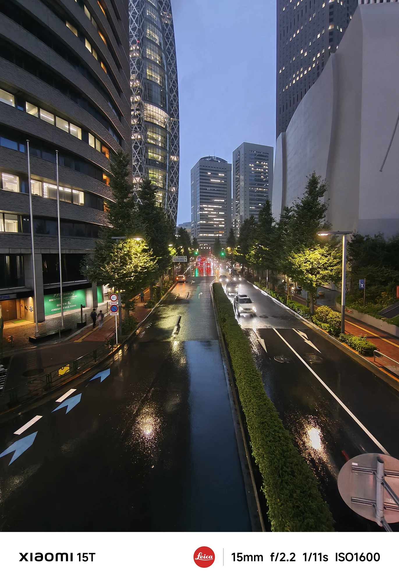 雨に濡れた夜の交差点。両側の高層ビルと奥のタワーがライトアップされ、傘をさした数人が写っている。撮影データ: 23mm, f/1.7, 1/27s, ISO800。