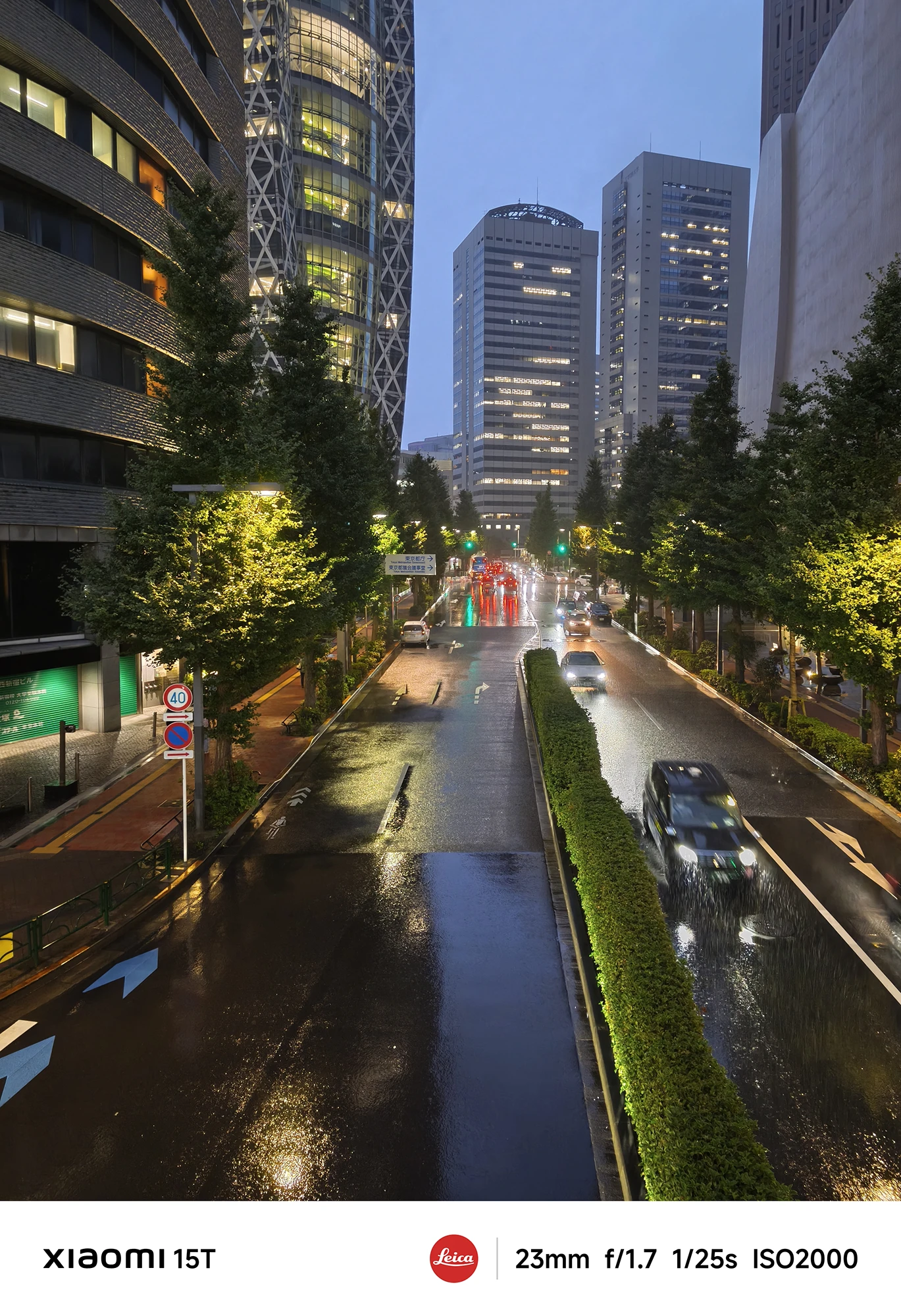 暗い雨の夜の都市の道路。左右に高層ビルが立ち並び、街路樹の緑が街灯に照らされている。濡れた路面に車のヘッドライトやテールランプが反射している。撮影データ: 23mm, f/1.7, 1/25s, ISO2000。