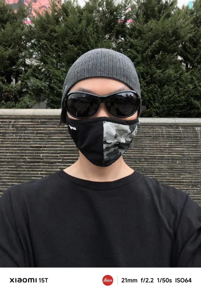 ニット帽、黒いサングラス、迷彩柄のマスクを着用し、黒いTシャツを着た人物のアップの自撮り写真。背景は濃い緑の生け垣と石垣のような壁。Xiaomi 15T撮影データ: 21mm, f/2.2, 1/50s, ISO64。