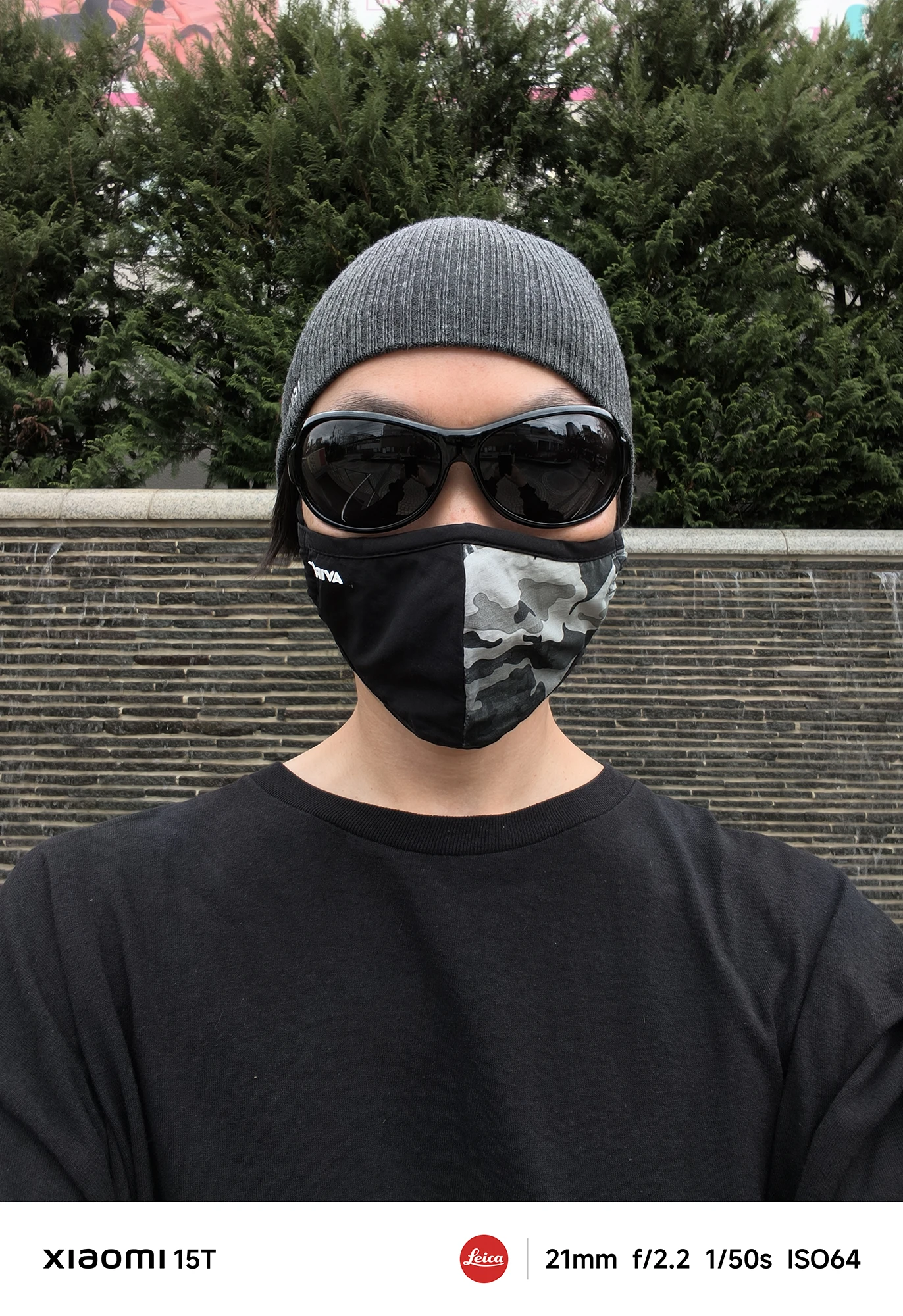 ニット帽、黒いサングラス、迷彩柄のマスクを着用し、黒いTシャツを着た人物のアップの自撮り写真。背景は濃い緑の生け垣と石垣のような壁。撮影データ: 21mm, f/2.2, 1/50s, ISO64。