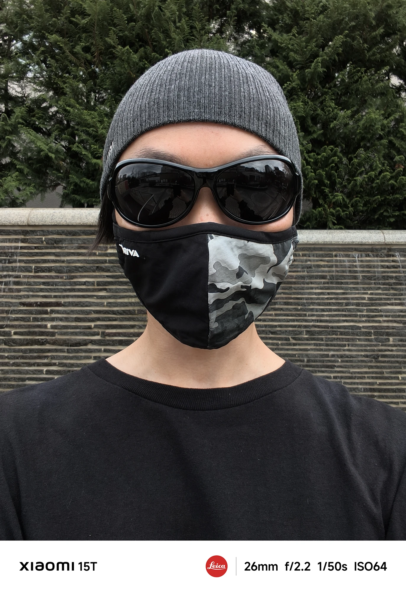 ニット帽、黒いサングラス、迷彩柄のマスクを着用し、黒いTシャツを着た人物のアップの自撮り写真。背景は濃い緑の生け垣と石垣のような壁。撮影データ: 26mm, f/2.2, 1/50s, ISO64。