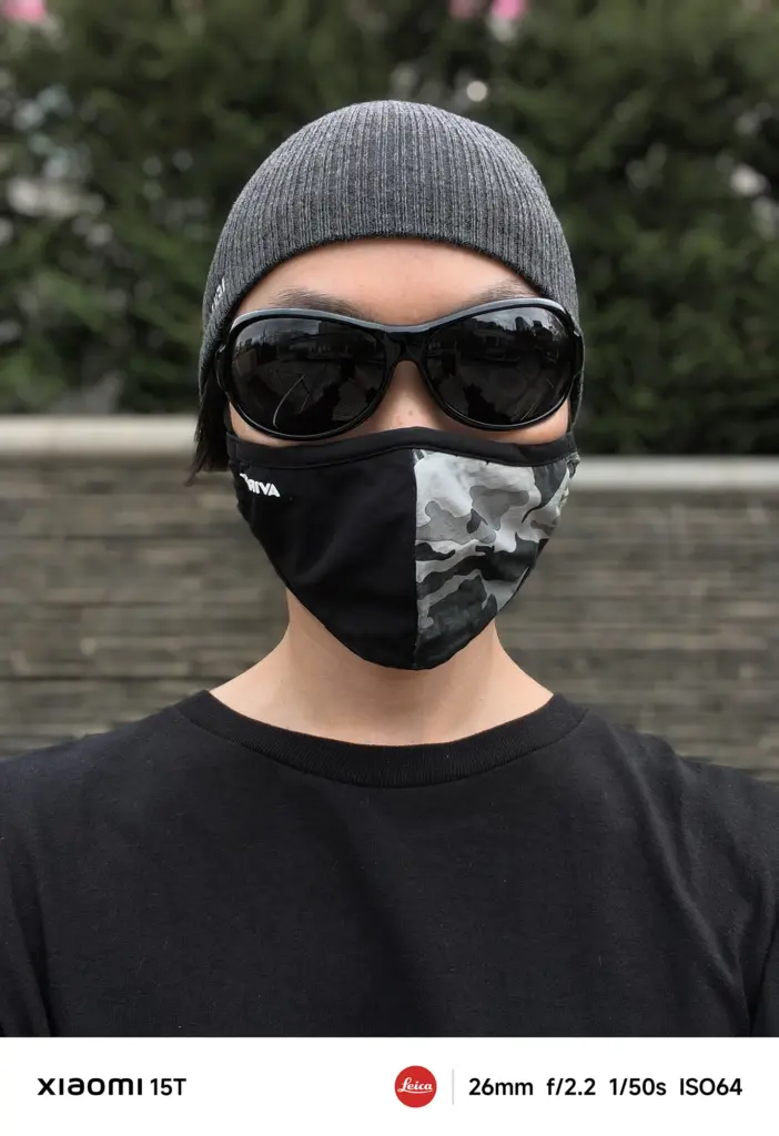 ニット帽、黒いサングラス、迷彩柄のマスクを着用した人物のポートレート写真。背景の生け垣と石垣がボケており、人物が強調されている。Xiaomi 15T撮影データ: 26mm, f/2.2, 1/50s, ISO64。