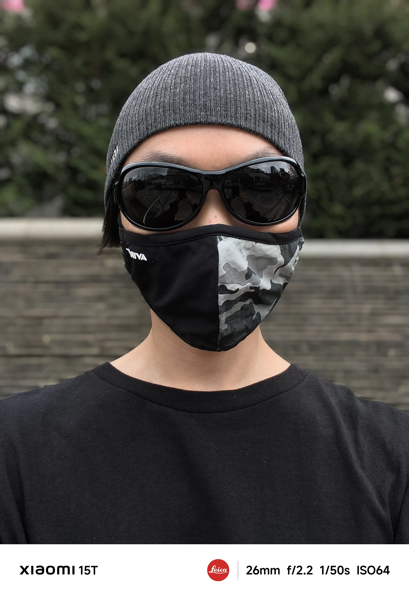 ニット帽、黒いサングラス、迷彩柄のマスクを着用した人物のポートレート写真。背景の生け垣と石垣がボケており、人物が強調されている。撮影データ: 26mm, f/2.2, 1/50s, ISO64。