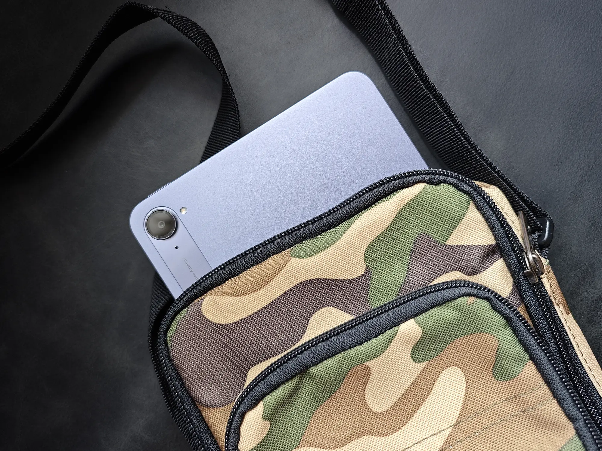 パープル/ブルーグレー系のコンパクトなタブレットを、カモフラージュ(迷彩)柄のショルダーバッグに収納している様子。携帯性の高さとカメラ部分が見えている。