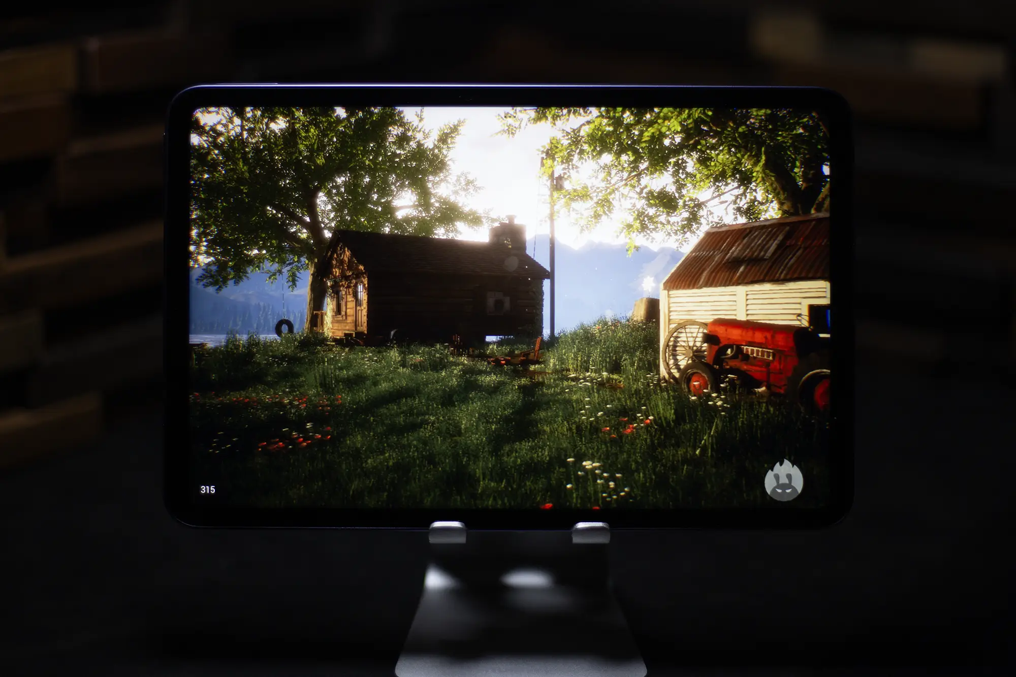 タブレット端末でゲームをプレイする様子。画面には、リアルなグラフィックで丸太小屋や赤いトラクターが描かれた、田園風景のゲーム画面が表示されている。