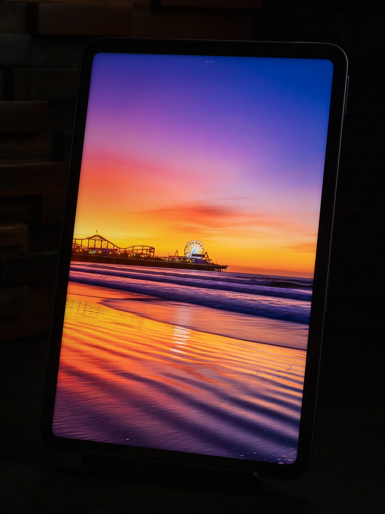 タブレットのディスプレイに表示された、ビーチと桟橋の鮮やかな夕焼け(日の出)写真。観覧車とオレンジ、紫、青のグラデーションの空が写っている。