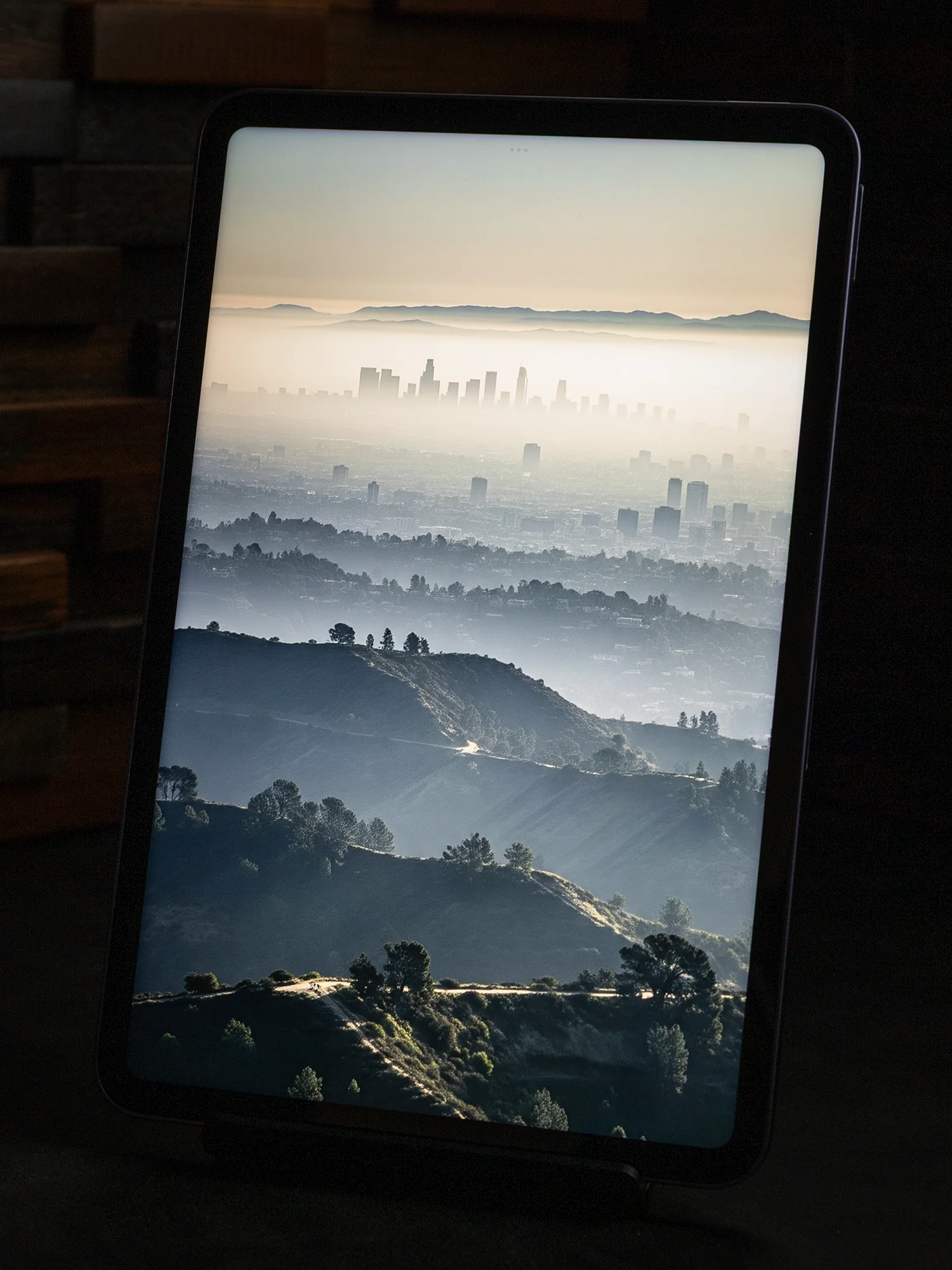 タブレットのディスプレイに表示された、霧(霞)のかかった遠景の都市風景写真。手前の丘陵地帯と、奥の山並みの手前に高層ビル群のシルエットが浮かび上がっている。