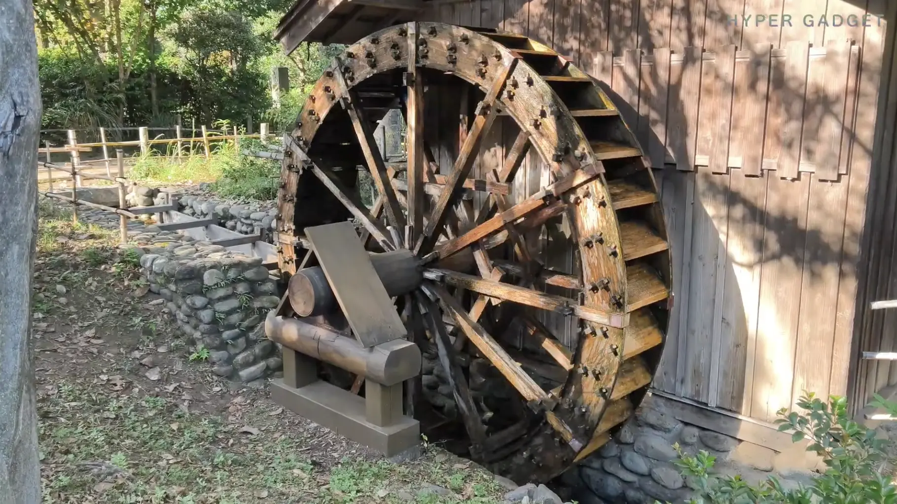 木造の建物と水路の横に設置された大きな木製水車（ウォーターホイール）。水車の一部が水に濡れており、周囲には石垣と草が生えている。