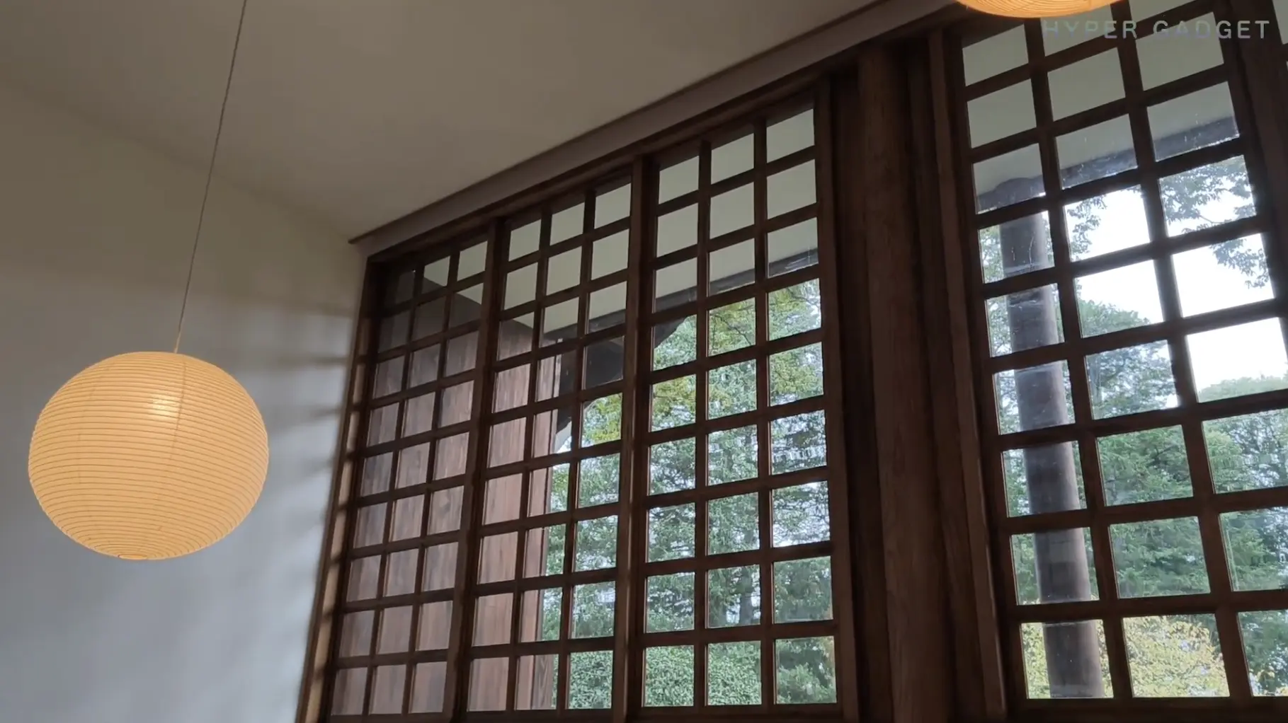 和風建築の室内から見た景色。天井まで届く大きな木枠の格子窓が印象的で、窓の外には緑の木々が見える。左側には和紙のような素材の丸いペンダントライトが吊るされている。