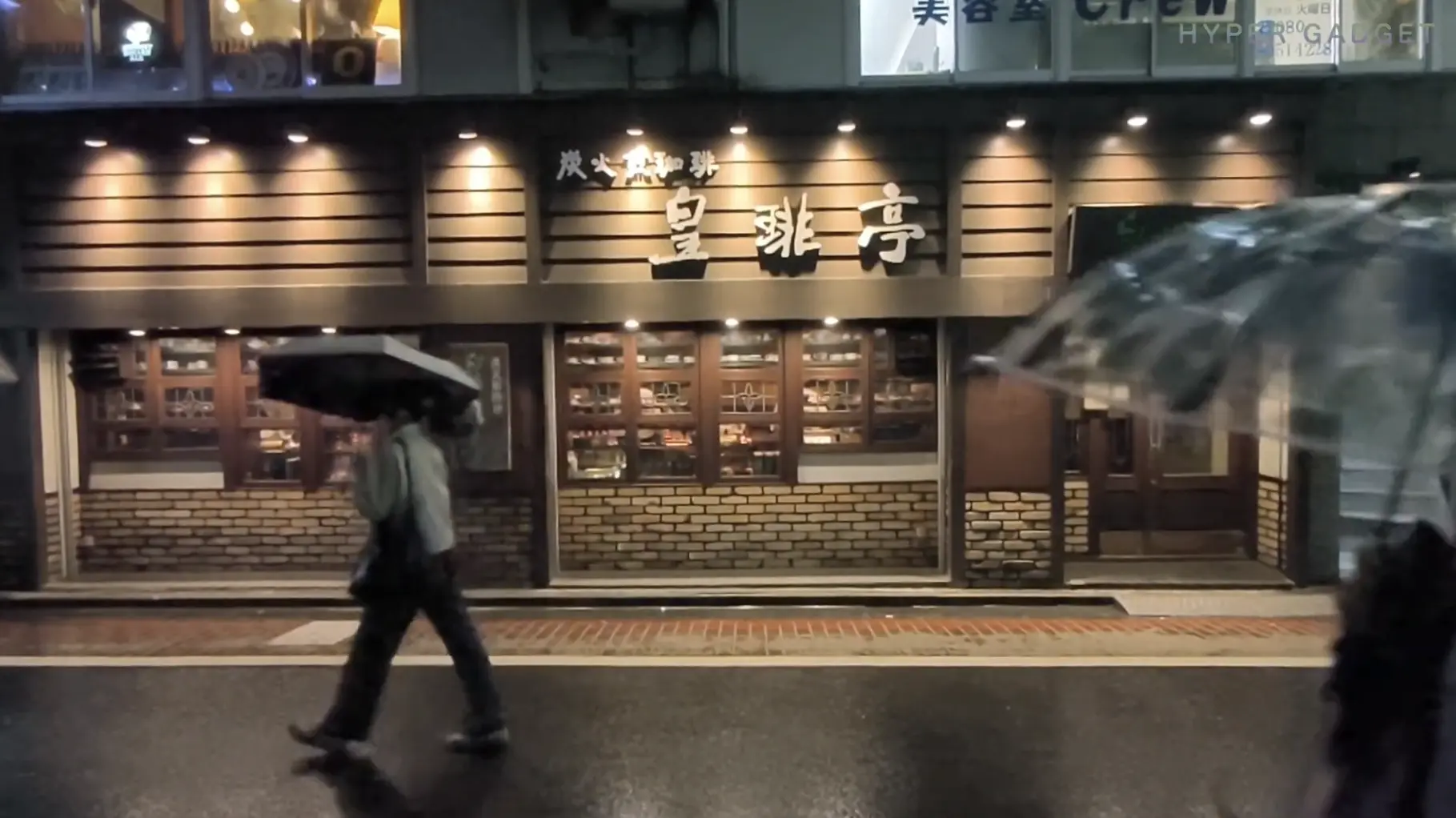 雨の夜の街角にある喫茶店またはコーヒーショップ。木目とレンガ調の外観で、「炭火焙煎 珈琲亭」と書かれた看板が光っている。傘をさした人物が手前の濡れた歩道を歩いている。