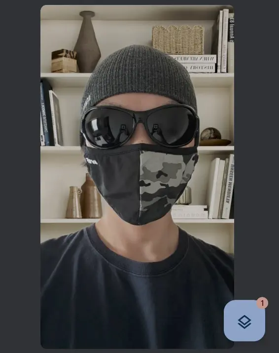 Google Meetのビデオ通話画面のスクリーンショット。ニット帽、黒いサングラス、迷彩柄のマスクを着用した人物の顔が映っている。背景は本棚で、本や花瓶などの小物が置かれている。