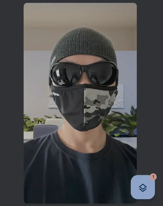 Google Meetのビデオ通話画面のスクリーンショット。ニット帽、黒いサングラス、迷彩柄のマスクを着用した人物の顔が映っている。背景はぼかしや合成されたグリーン系の観葉植物と明るい壁。
