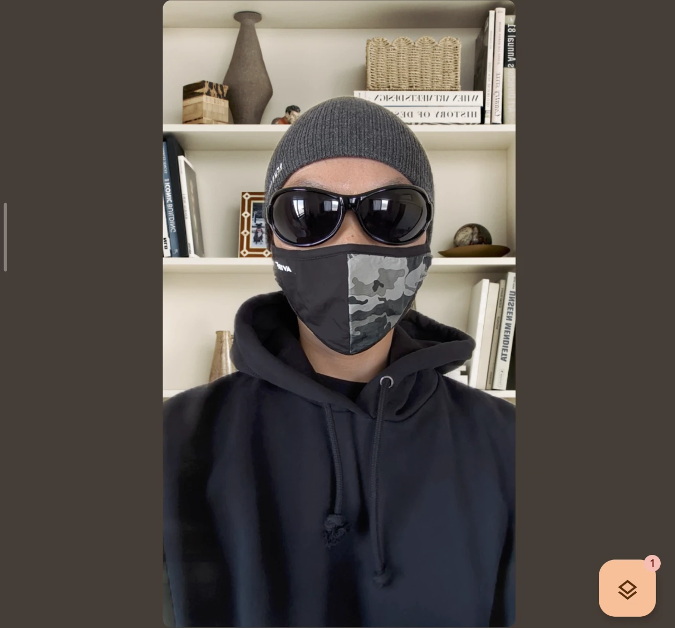ビデオ通話画面(またはポートレートモード)で撮影された、サングラスと迷彩マスクを着用した人物の正面写真。背景の本棚が人工的にボケて強調されている。