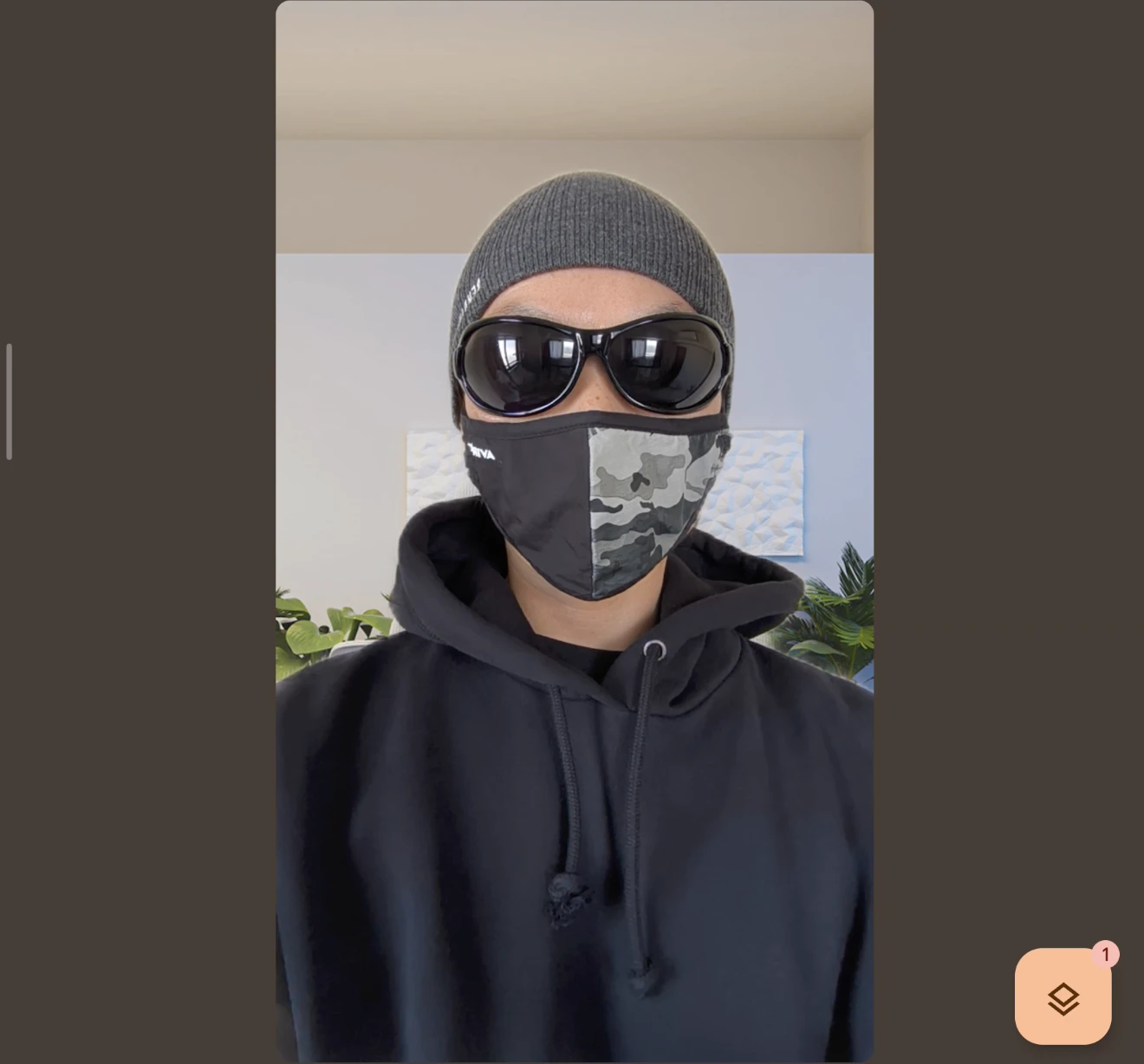 ビデオ通話画面(またはポートレートモード)で撮影された、サングラスと迷彩マスクを着用した人物の正面写真。背景の室内にある植物が人工的にボケて強調されている。