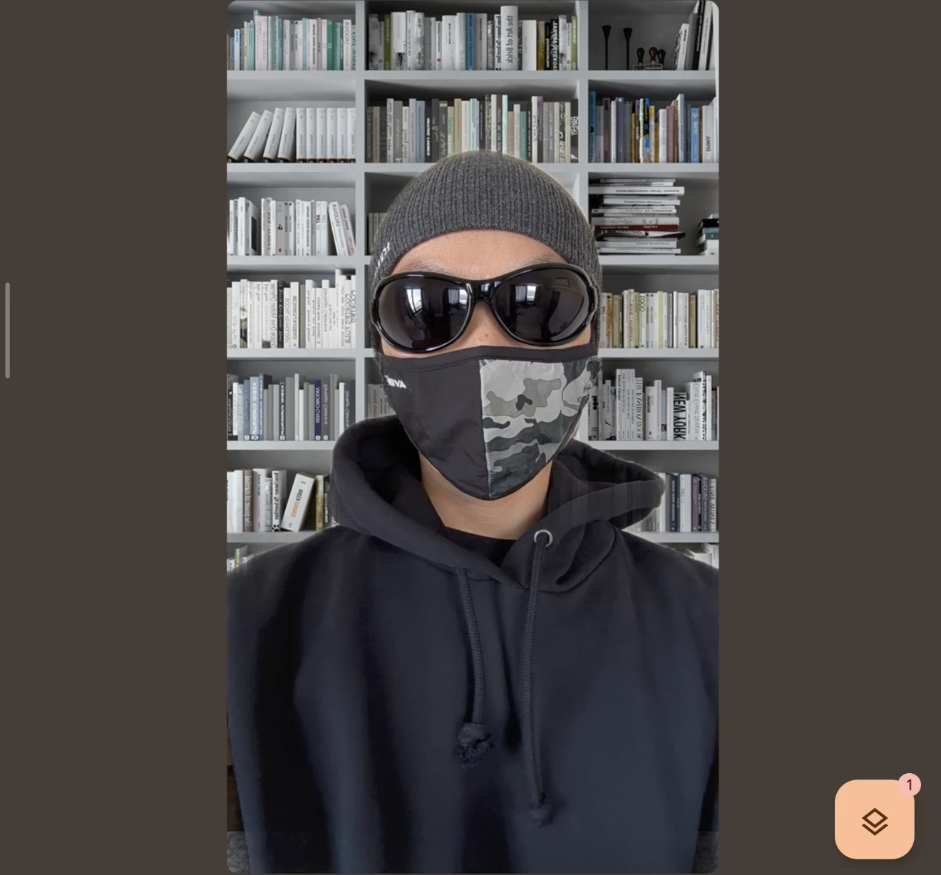 ビデオ通話画面(またはカメラアプリ)で撮影された、サングラスと迷彩マスクを着用した人物の正面写真。背景が本が並んだ本棚の画像に合成(置き換え)されている。