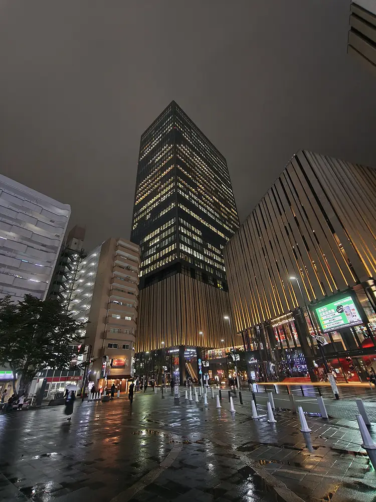 夜の雨上がりの都市風景写真。下から見上げるような構図で、高層ビルと商業施設、濡れた路面の光の反射がダイナミックに写っている。