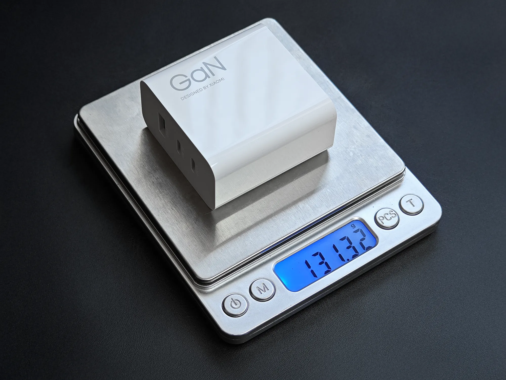 Xiaomi製の白いGaN（窒化ガリウム）充電アダプターをデジタルスケールに載せて重量を計測している写真。スケールのディスプレイには、充電器の重量が131.3グラムと表示されている。充電器本体には「GaN DESIGNED BY XIAOMI」と印字されている。